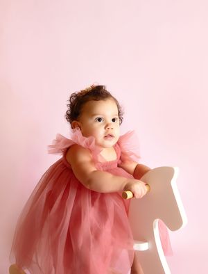 Baby girl in pink tulle dress leaning on wooden rocking horse against pink background.