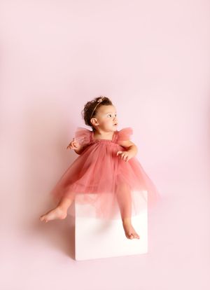 Baby girl in pink tulle dress sits on white cube against pink background.