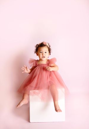 Baby girl in pink tulle dress and floral crown sits on white cube against soft pink background.
