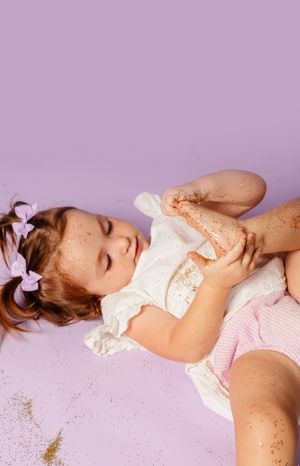 A young girl with purple bows lies on a lavender background, wearing a white dress, covered in gold glitter.