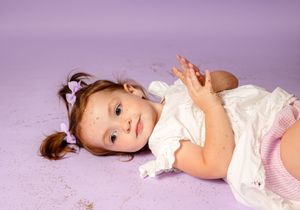 Toddler girl in white dress lying on purple floor, looking up with hand raised, wearing pigtails.