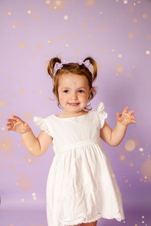 Cheerful toddler girl in white dress with pigtails posing against purple bokeh background.