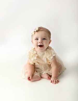 Smiling baby girl in cream lace floral romper and bow headband sitting on white background.