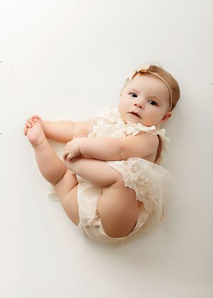 A cute baby girl in a cream lace romper and bow headband lying on a white background.