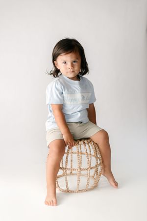 A young toddler sits on a woven rattan stool wearing a light blue tee and khaki shorts against a white background.
