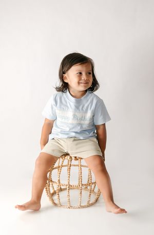 Toddler smiling in a light blue birthday tee and khaki shorts, sitting on a woven rattan stool against a white background.