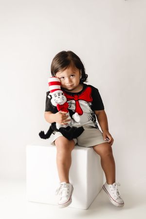 Young child sitting on white cube holding Cat in the Hat stuffed toy, wearing matching Dr. Seuss graphic tee and white sneakers.