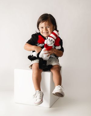 Smiling toddler sitting on white cube holding Cat in the Hat plush toy, wearing black shirt and denim shorts.
