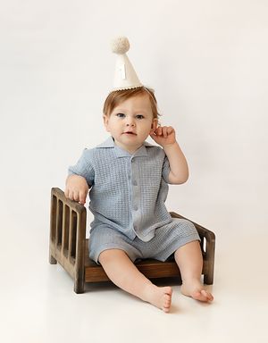 Baby boy in blue knit romper wearing a cream party hat sits on a wooden bench for his first birthday photo.