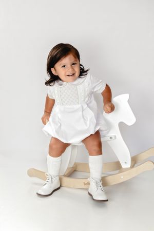 Smiling toddler in white dress and knee socks sitting on a wooden rocking horse against a white background.