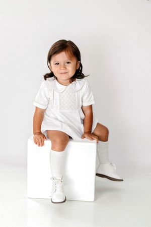 Toddler girl in white smocked dress and knee-high boots sits smiling on white cube in a bright studio.