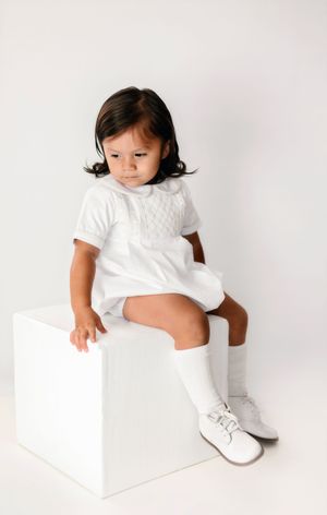 Toddler in white outfit and knee socks sits on white cube, wearing white shoes against a white background.