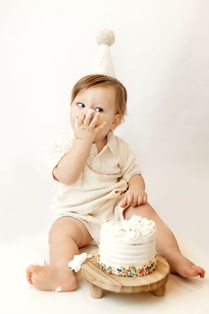 Baby in a white party hat smashing a sprinkle cake, licking frosting off hand during a first birthday cake smash.