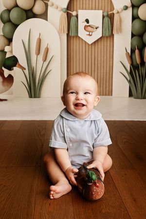 Smiling baby in blue romper sits on wood floor with duck-themed backdrop for first birthday cake smash photo.