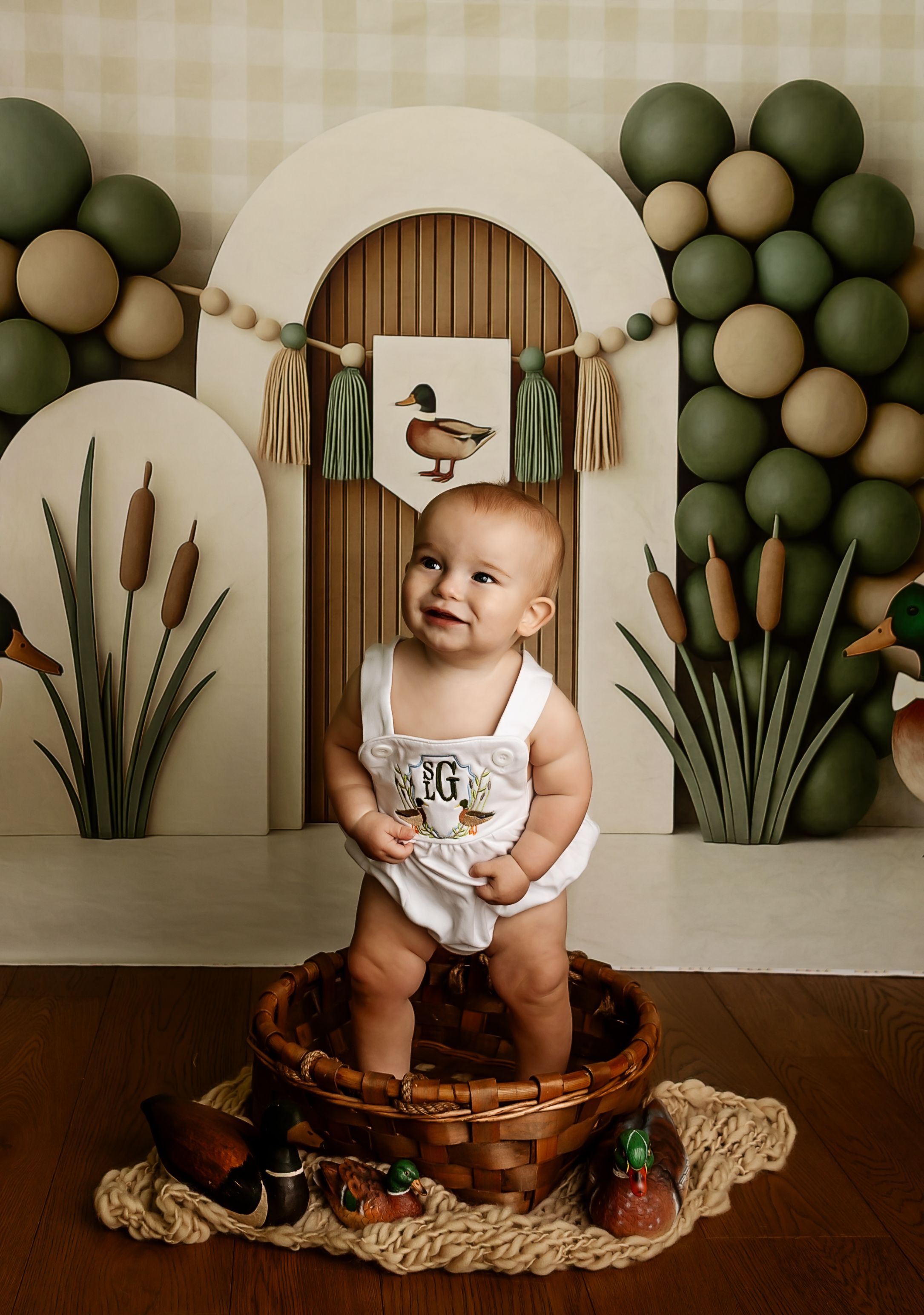 Baby in white romper sits in wicker basket, surrounded by duck-themed decor with cattails and green balloon backdrop.