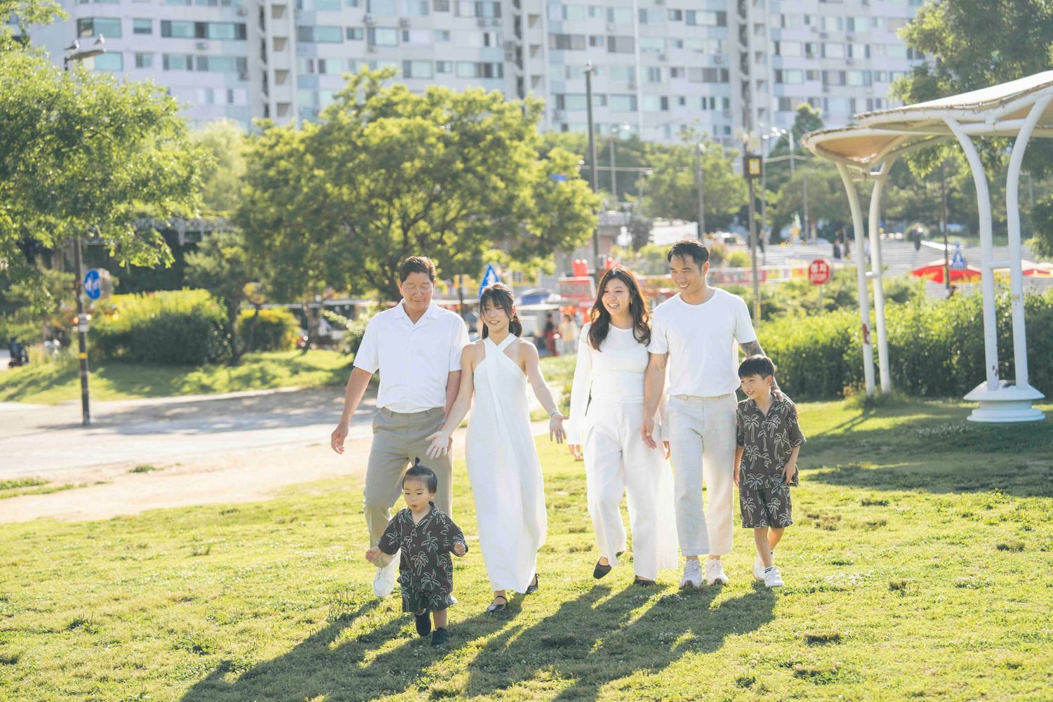 Happy Asian family walking together in a sunny green park, parents and children dressed in white and casual outfits.