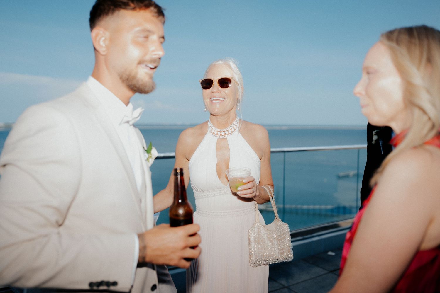 Three guests socializing at an outdoor rooftop wedding reception with ocean views on a sunny day.