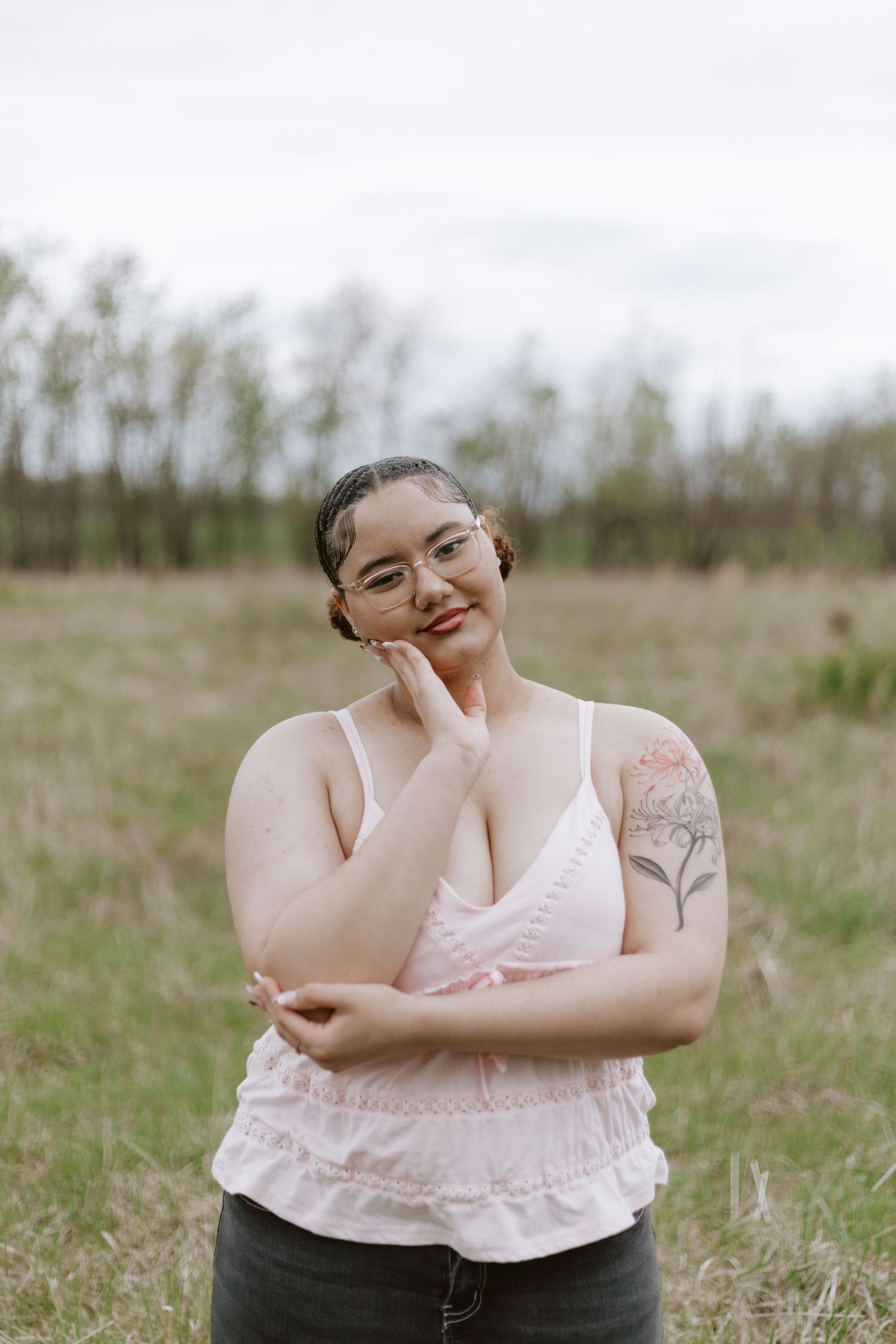 Plus-size woman with glasses and floral arm tattoo smiling in pink cami top, posing outdoors in a green field.