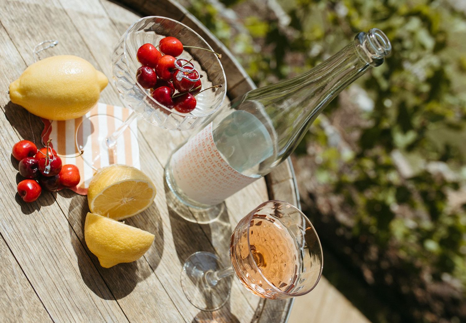 Sunlit outdoor table styled with rose wine, lemons, cherries, and a glass bottle for a summer picnic.