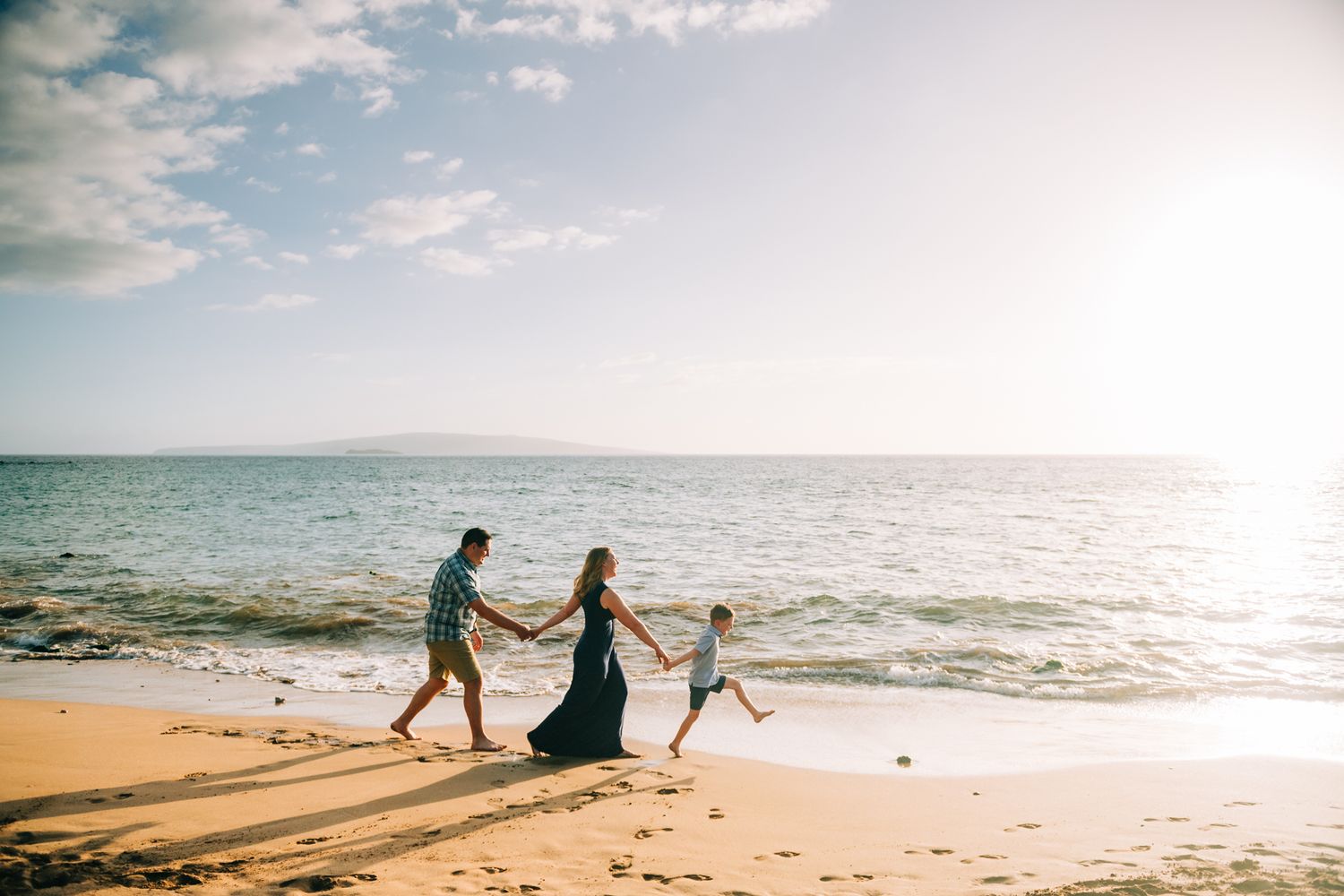 Family of three holding hands and playing on a sunny sandy beach with ocean waves and mountains in background.