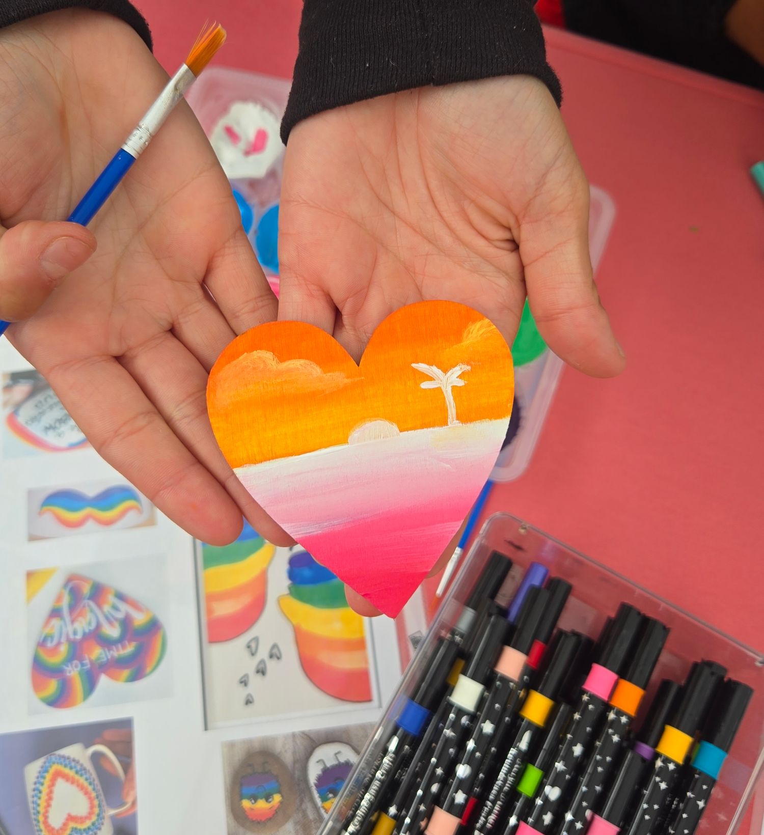 Hands holding a painted heart-shaped canvas with a tropical sunset scene, surrounded by colorful markers and art supplies.