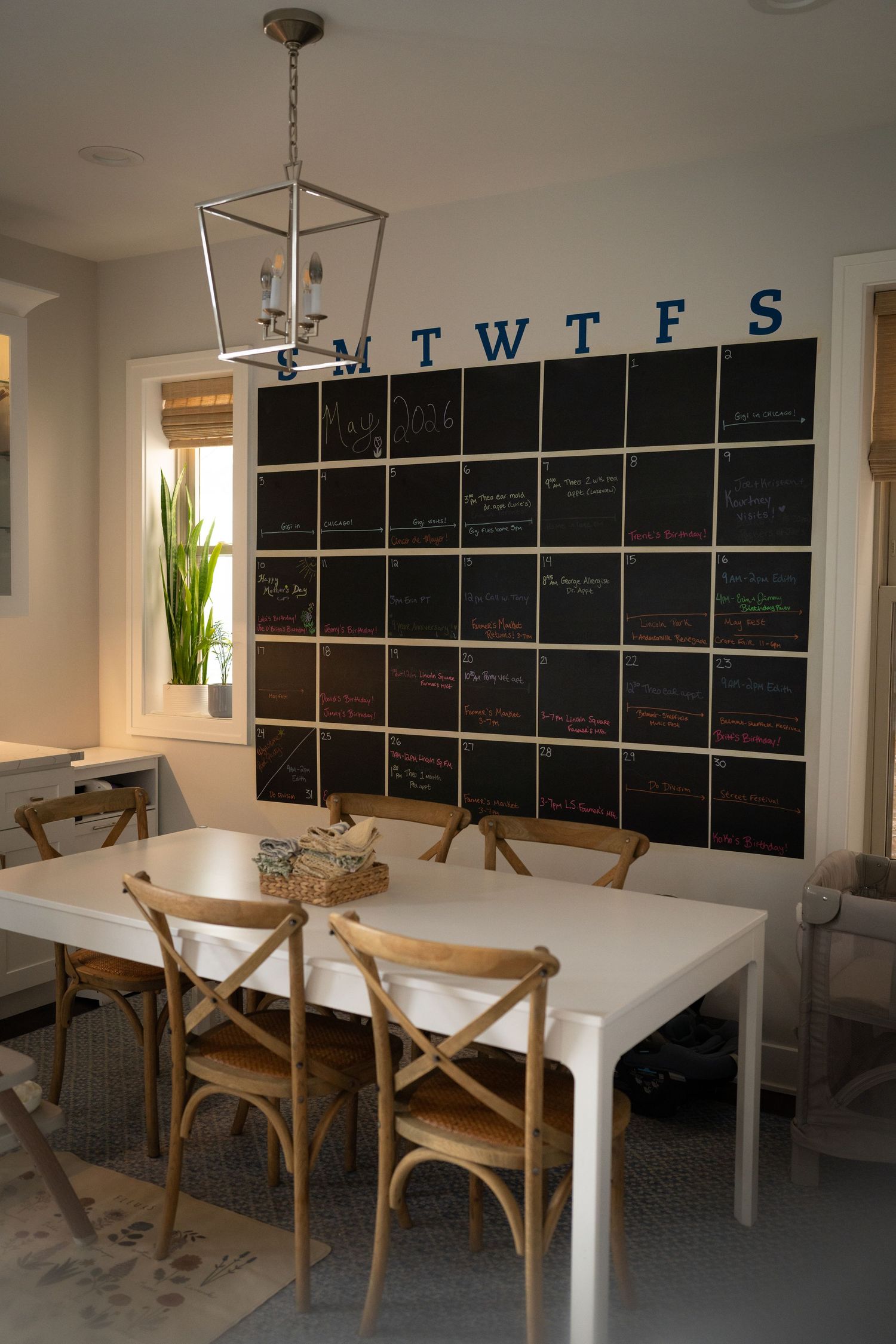 Dining room with white table, wooden chairs, and large chalkboard calendar wall with SMTWTFS labels and lantern pendant light.