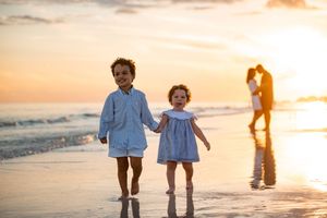 Two young children hold hands walking on a beach at sunset with their parents embracing in the background.