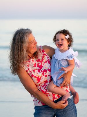 Grandmother holding laughing toddler girl on beach at sunset, both smiling joyfully near ocean waves.