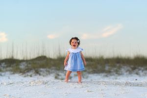 Toddler girl in blue dress laughing on white sand beach with sea oats and pastel sky backdrop.