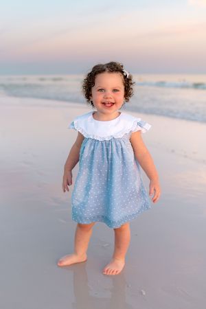 A smiling toddler girl in a blue polka dot dress stands barefoot on a sandy beach at sunset.