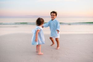 Two toddlers in light blue outfits playing barefoot on a sandy beach at sunset near gentle ocean waves.