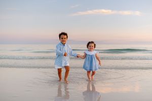 Two young children holding hands and smiling on a sandy beach at sunset, waves gently lapping behind them.