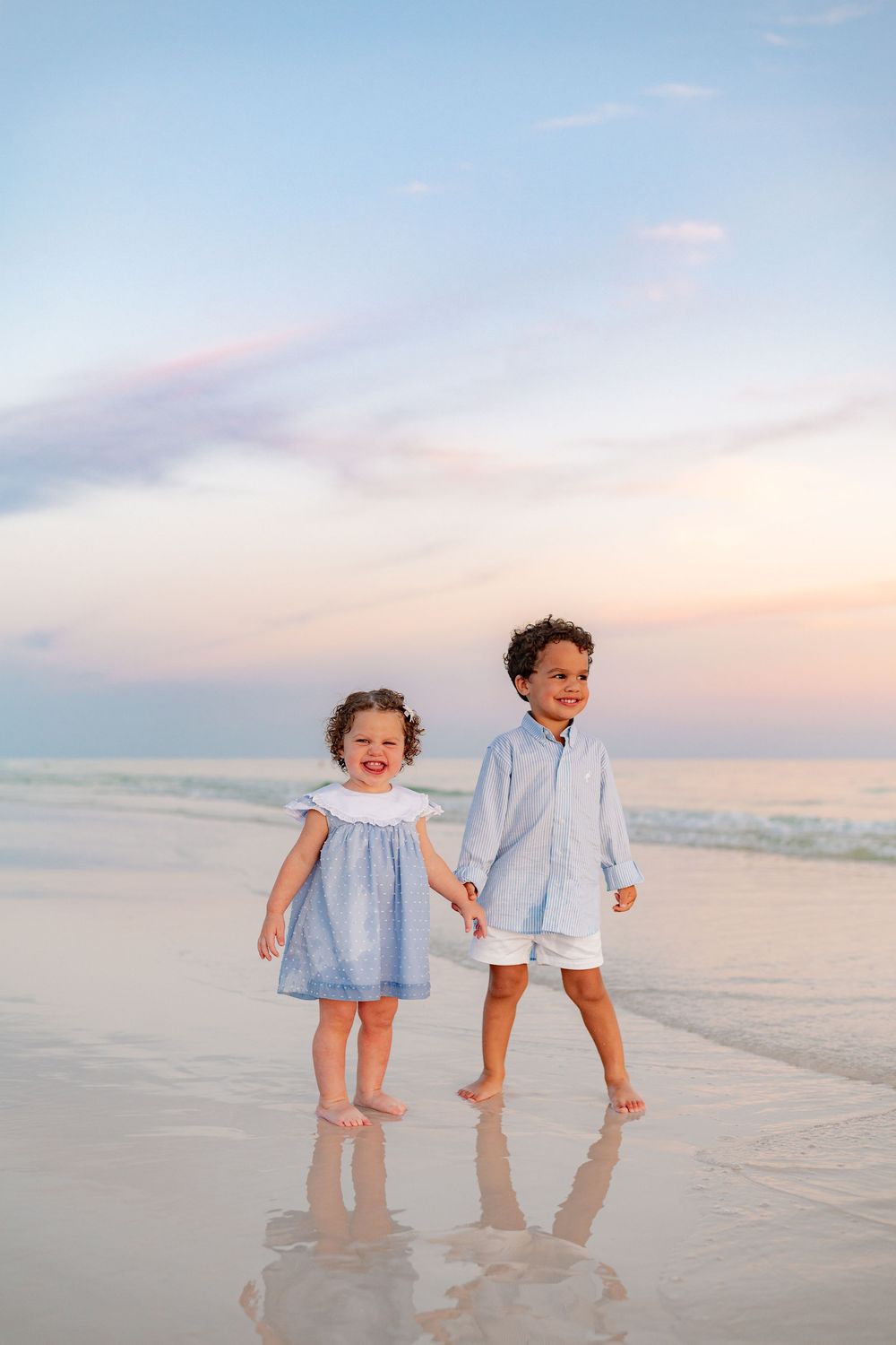 Two young children holding hands on a beach at sunset, wearing matching blue outfits, smiling and walking along the shoreline.