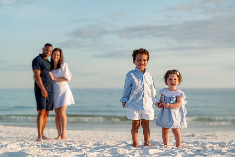 Family of four poses on a white sand beach, two young children smiling in the foreground, parents embracing behind them.
