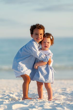 Two young siblings hug on a white sand beach at sunset, both wearing matching blue outfits.