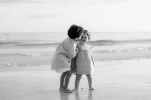Two young children share a tender moment on a sandy beach, with ocean waves in the background, in black and white.