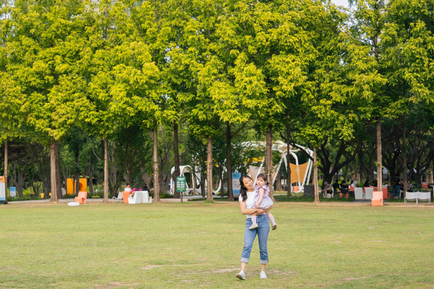 A woman holds a child in a lush green park surrounded by tall trees on a sunny day.