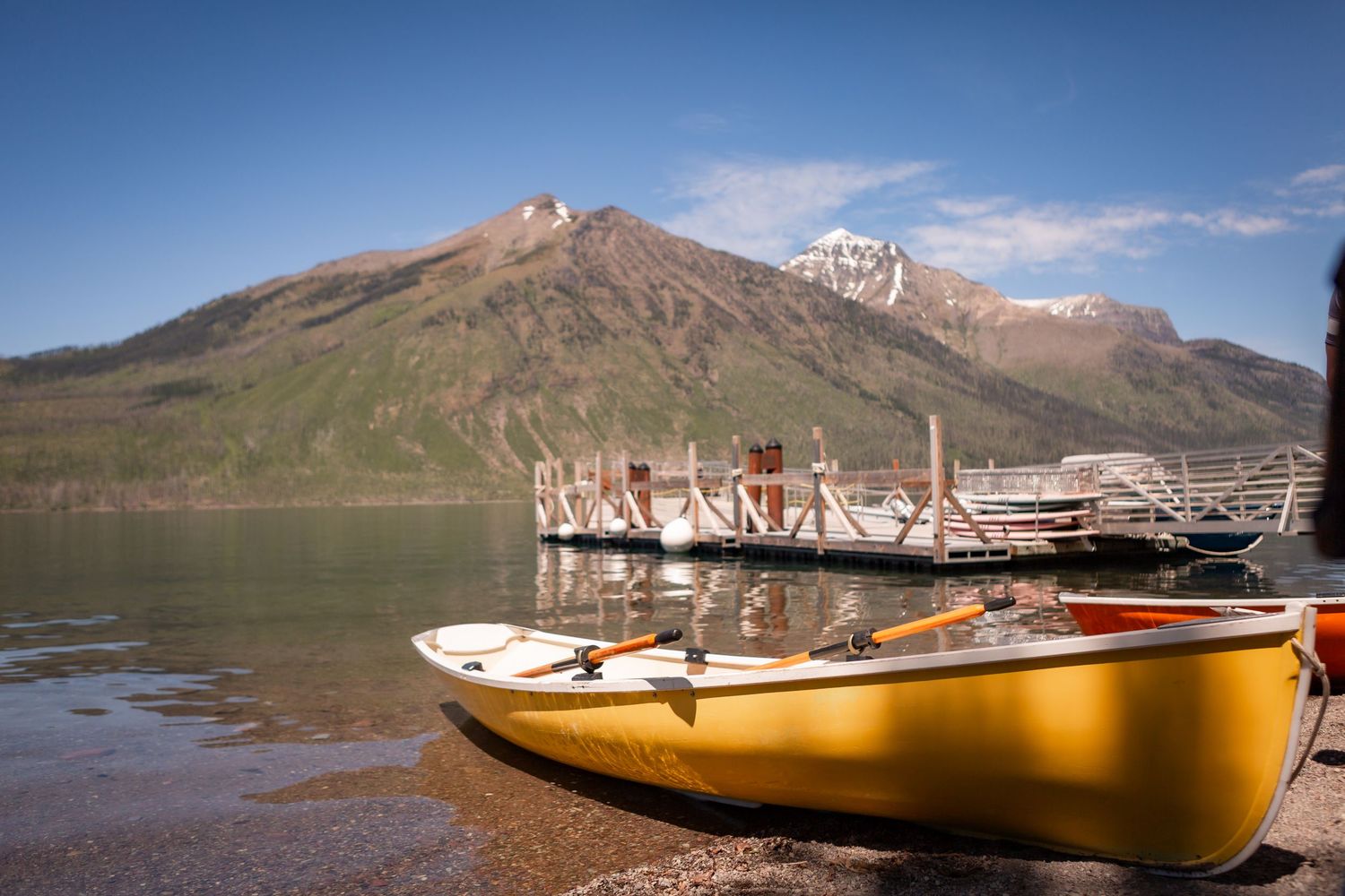 Yellow canoe on the shore of a calm mountain lake with a wooden dock and snow-capped peaks under blue sky.