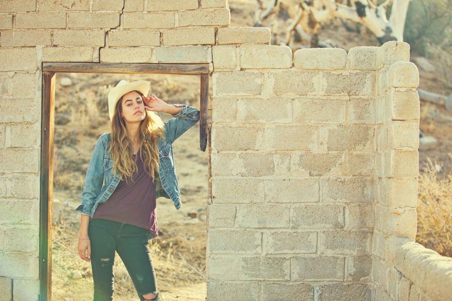 Young woman in denim jacket posing in rustic stone doorway, golden hour sunlight, bohemian fashion outdoor shoot.