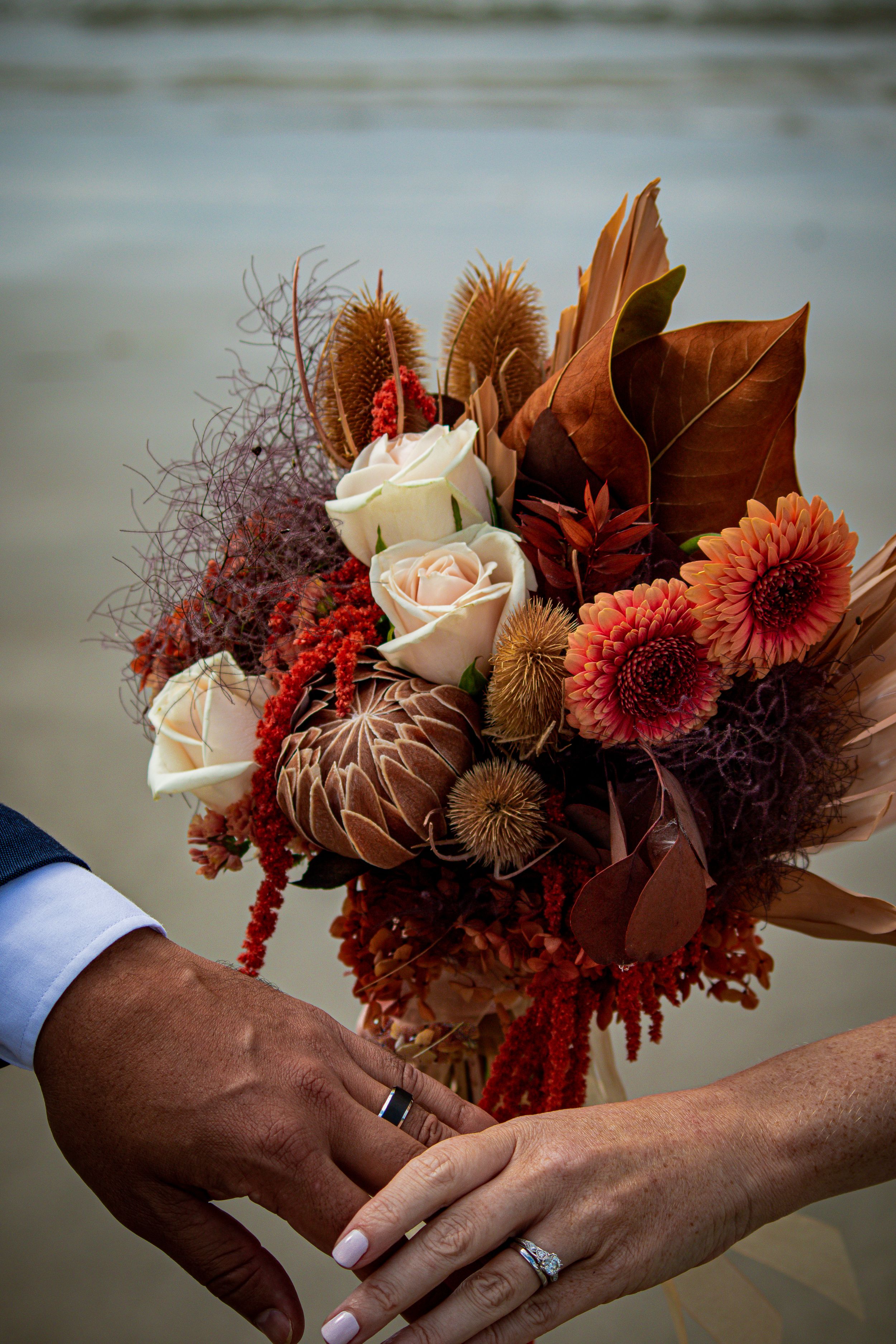Bride and groom holding an autumn wedding bouquet with roses and dahlias on a beach.