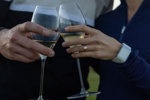Couple clinking white wine glasses in a toast, woman wearing a diamond engagement ring.