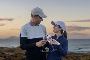 Couple toasting with wine glasses at sunset by the ocean, both wearing golf attire and caps.
