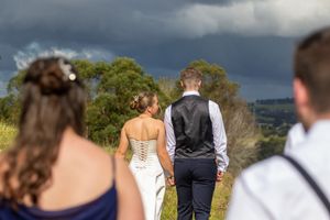 Bride and groom walking hand in hand through a field with stormy skies and rolling hills behind.