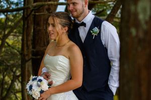 Bride holding blue and white bouquet stands with groom in navy vest among tall pine trees.