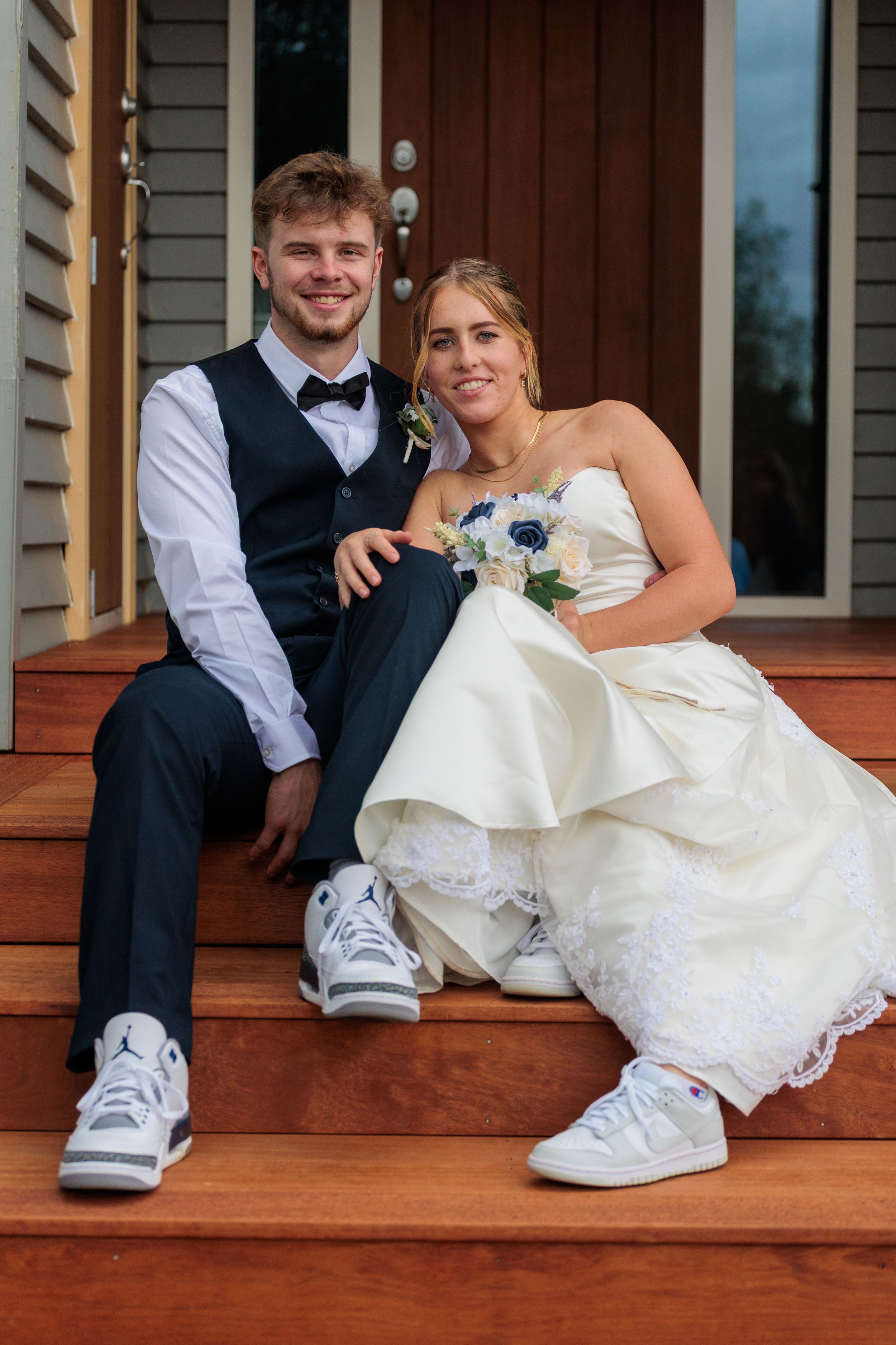 Bride and groom sitting on wooden porch steps wearing white sneakers, smiling at the camera.
