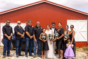 Wedding party group photo in front of a red barn with bride in white and guests in black attire.