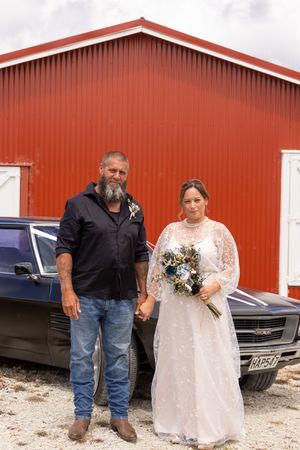 Bride and groom standing in front of a red barn beside a classic black car on their wedding day.
