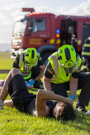 Airport rescue firefighters in high-visibility gear providing first aid to an injured person on grass.