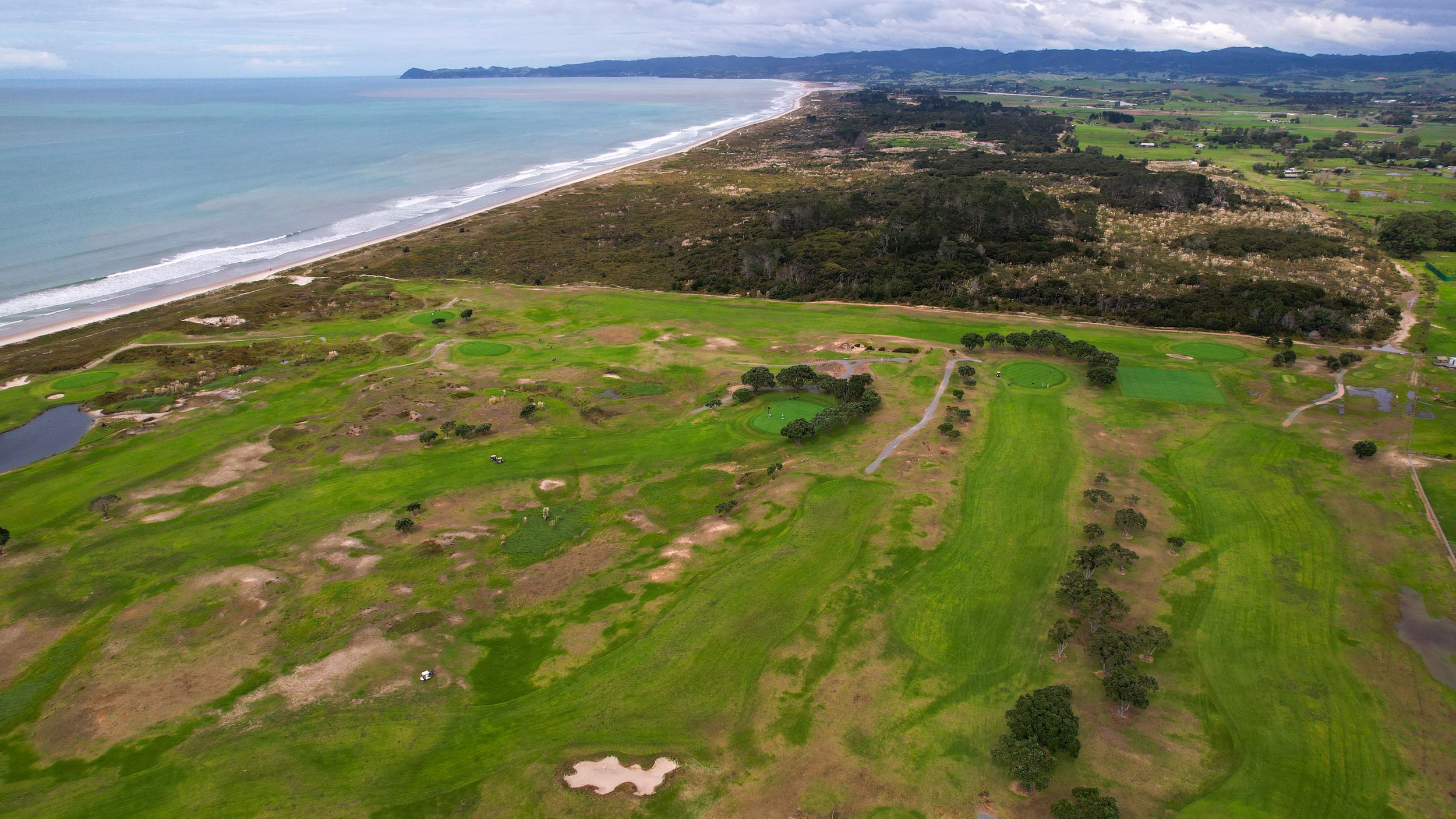 Aerial view of a coastal golf course with sandy dunes and ocean backdrop.