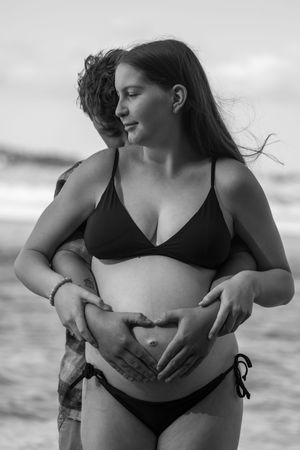 Black and white photo of a couple at the beach cradling a pregnant belly in a heart shape.